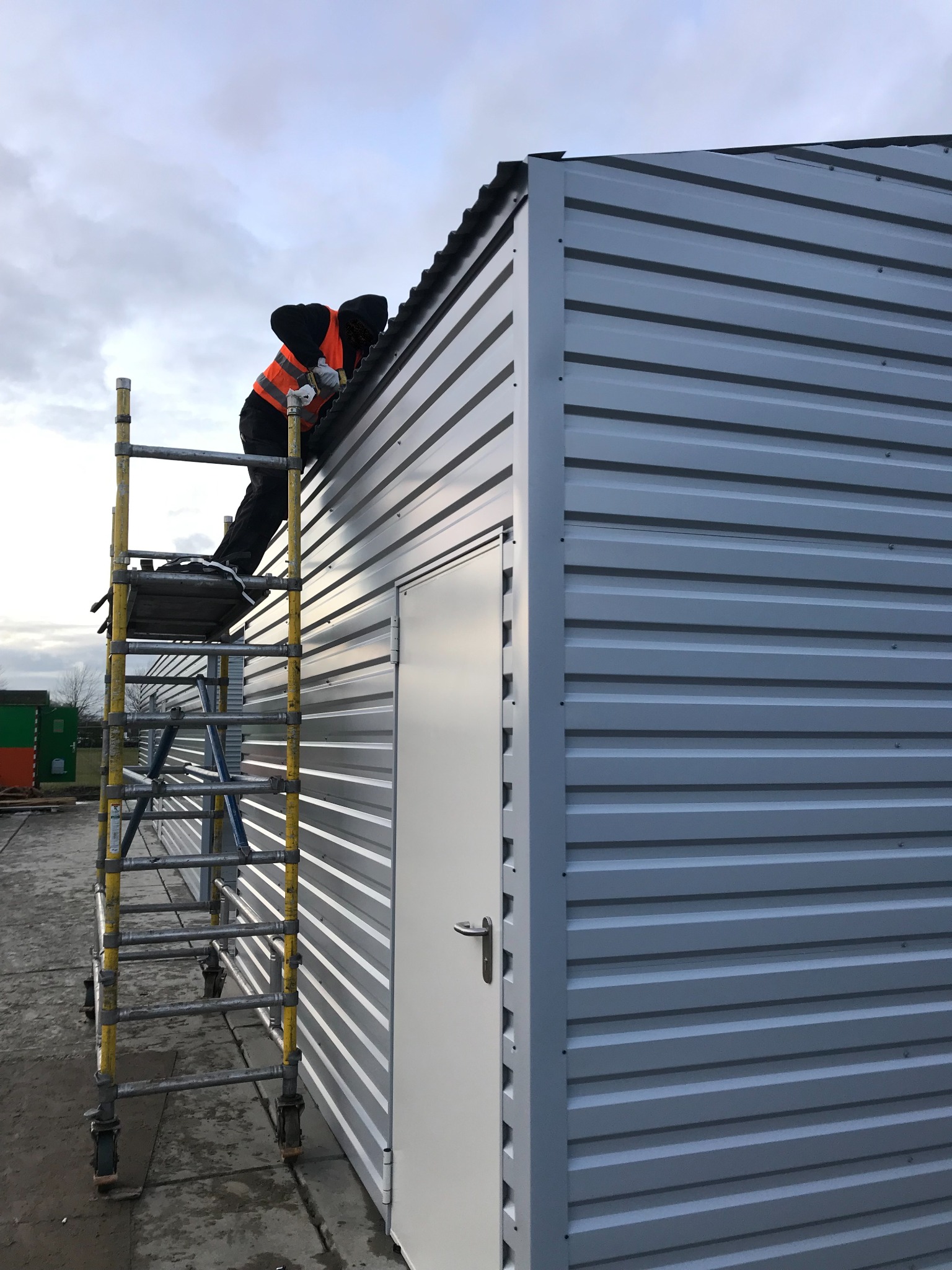 Pracownik w kamizelce odblaskowej na rusztowaniu montuje dach na budynku z szarej blachy trapezowej z białą furtką.