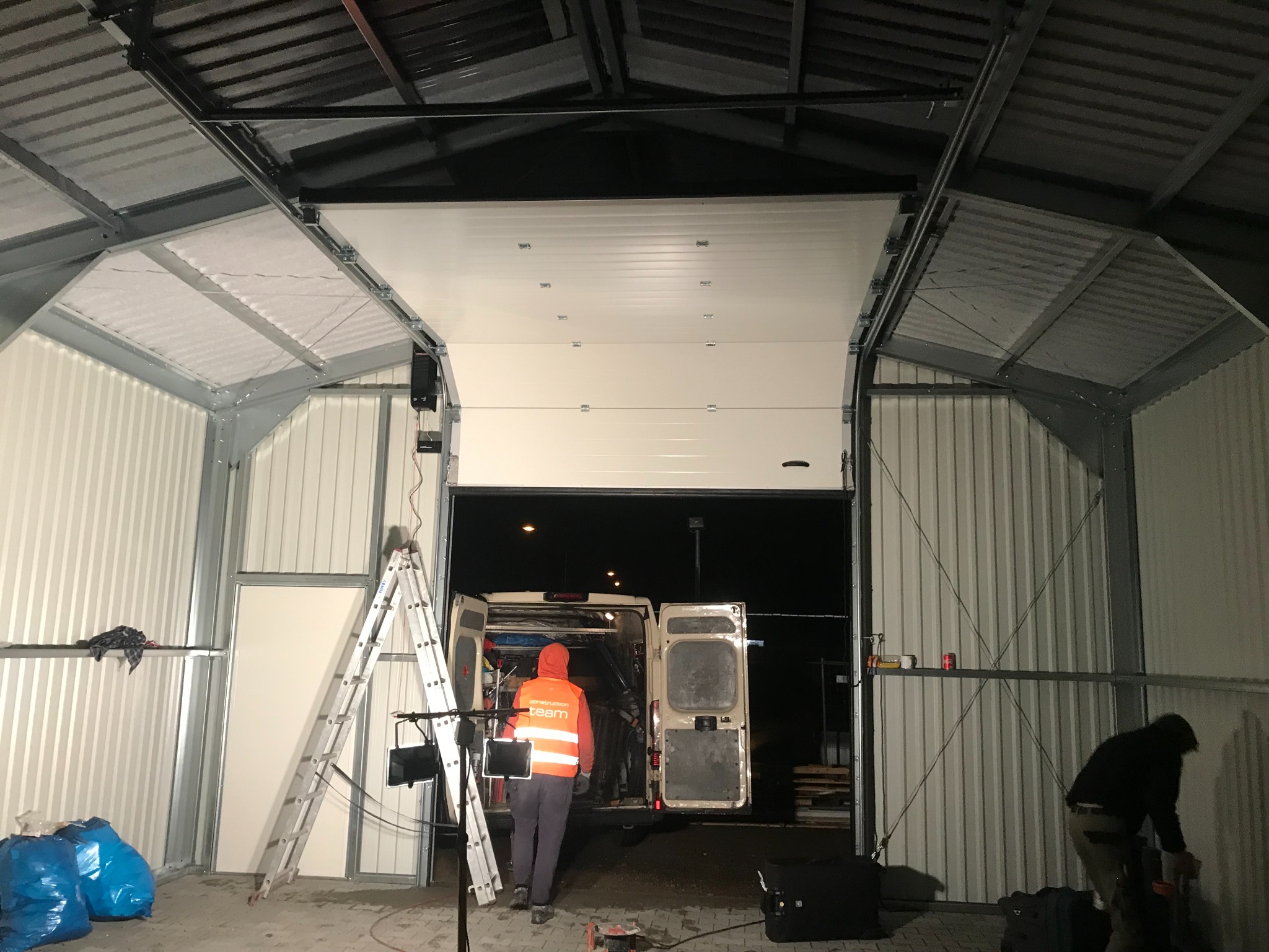 Wnętrze hali z częściowo otwartą białą bramą segmentową, widoczny pracownik w pomarańczowej kamizelce odblaskowej i bus z otwartymi drzwiami, drabina oparta o ścianę.