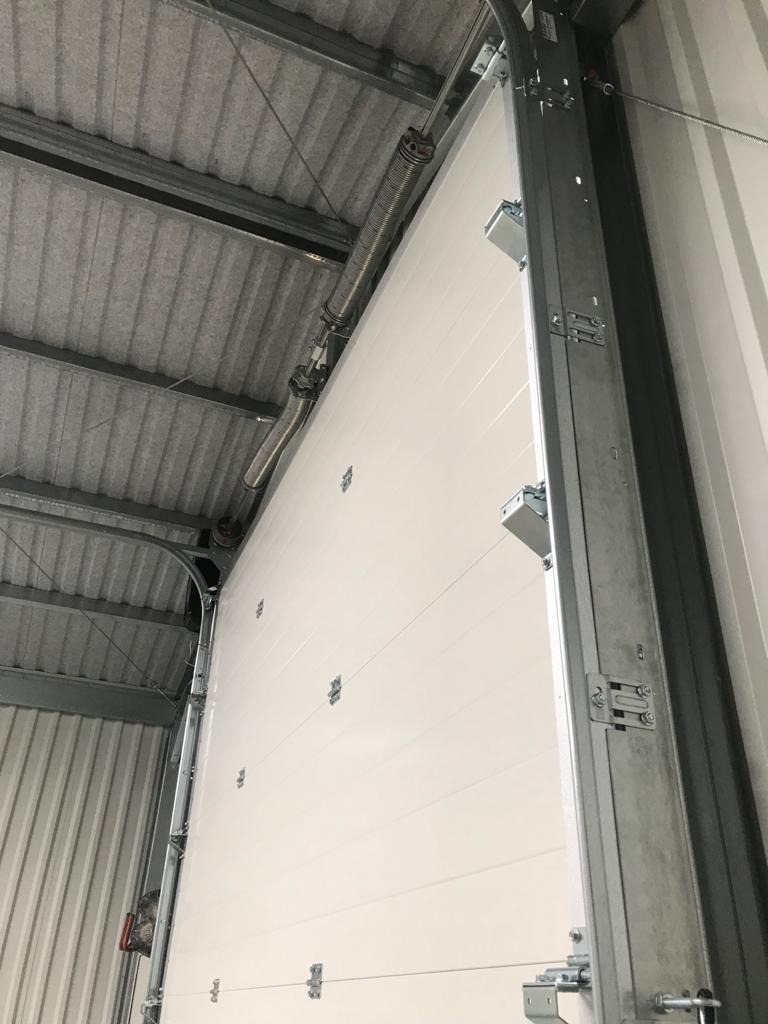 Wnętrze hali z widoczną białą segmentową bramą przemysłową w trakcie montażu, z widocznymi elementami konstrukcyjnymi i sprężynami nad bramą.
