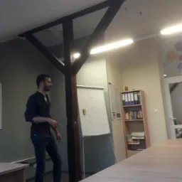 Mężczyzna gestykuluje podczas prezentacji przy tablicy w przestronnym biurze z drewnianymi elementami konstrukcyjnymi, w tle regał z segregatorami i przeszklone drzwi.