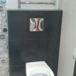 Zamontowana toaleta podwieszana z czarną obudową z płytek, chromowany przycisk spłukiwania i grzejnik drabinkowy w wykończonej łazience z białymi płytkami i dekoracyjnym pasem z wzorem geometrycznym.