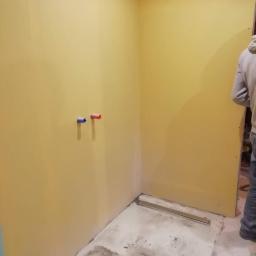 CLEAN MASTER BY TRUST TREE - Żółte ściany w trakcie prac wykończeniowych w łazience, widoczne rury doprowadzające wodę i odpływ liniowy w podłodze, częściowo przygotowanej pod płytki.