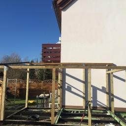 CLEAN MASTER BY TRUST TREE - Konstrukcja drewnianej pergoli w trakcie budowy przy białej ścianie budynku, widoczne słupy, belki, metalowe łączniki, narzędzia i materiały budowlane na ziemi.