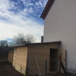 Nowo wybudowana drewniana szopa ogrodowa z otwartymi drzwiami, przylegająca do ściany domu, widoczna siatka ogrodzeniowa i dekoracyjny płotek.