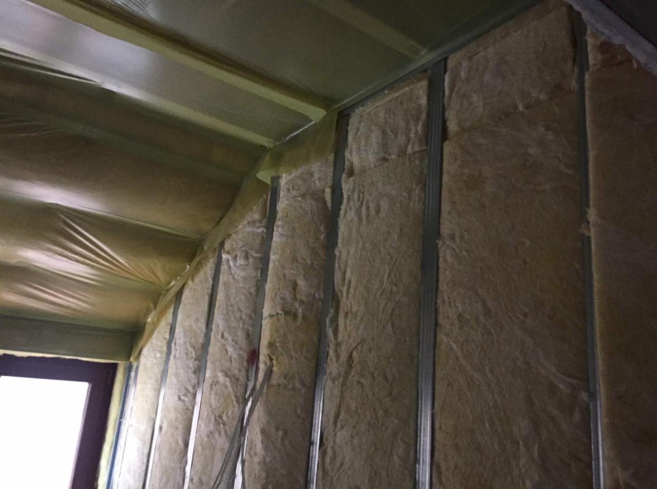 Wnętrze w trakcie prac wykończeniowych: metalowy szkielet konstrukcji, wypełniony wełną mineralną, przykryty folią paroizolacyjną na suficie, widoczny fragment okna.