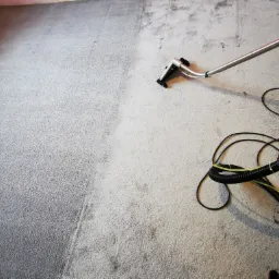 Dywan z wyraźną różnicą w czystości: lewa strona ciemniejsza, prawa jaśniejsza po czyszczeniu, widoczna dysza urządzenia do czyszczenia i węże.