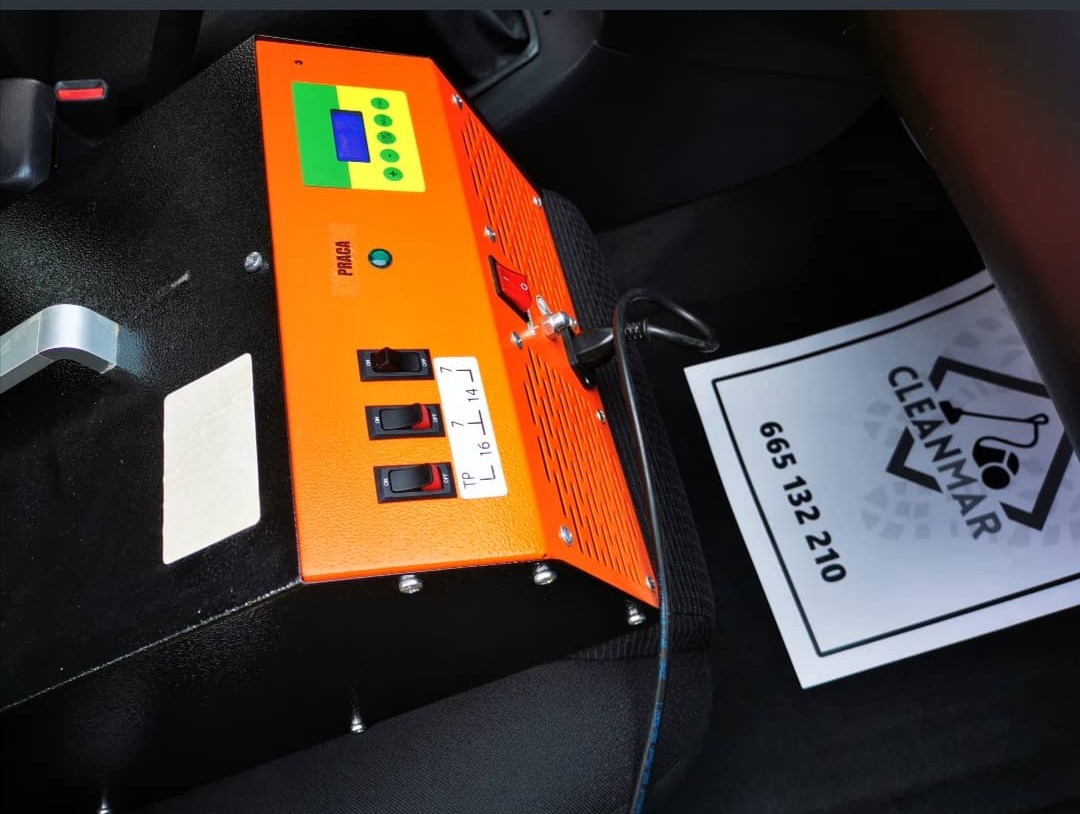 Pomarańczowo-czarne urządzenie z przełącznikami i wyświetlaczem, umieszczone na siedzeniu w samochodzie, obok ulotki z logo Cleanmar i numerem telefonu.