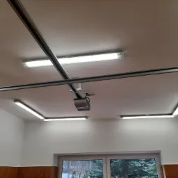 Wnętrze garażu z zamontowanymi lampami LED w kształcie litery U, połączonymi metalowymi szynami. Centralnie umieszczony automat do bramy garażowej.
