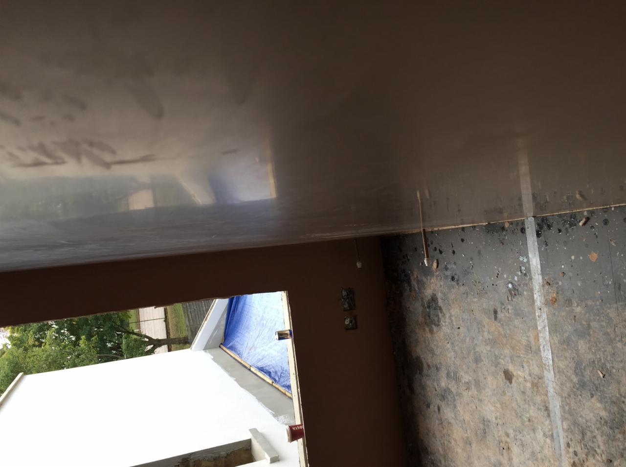 Wnętrze w trakcie prac wykończeniowych: gładka, jasna powierzchnia sufitu, brązowe ściany, widoczne przewody, surowa podłoga z plamami farby i widok na zewnątrz przez okno.