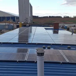 Instalacja paneli słonecznych na dachu przemysłowego budynku, widoczne odblaski nieba na panelach, w tle zabudowania gospodarcze i silosy.