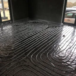 Instalacja ogrzewania podłogowego w surowym pomieszczeniu, widoczne rury grzewcze ułożone na folii i siatce, duże okna z widokiem na zewnątrz.