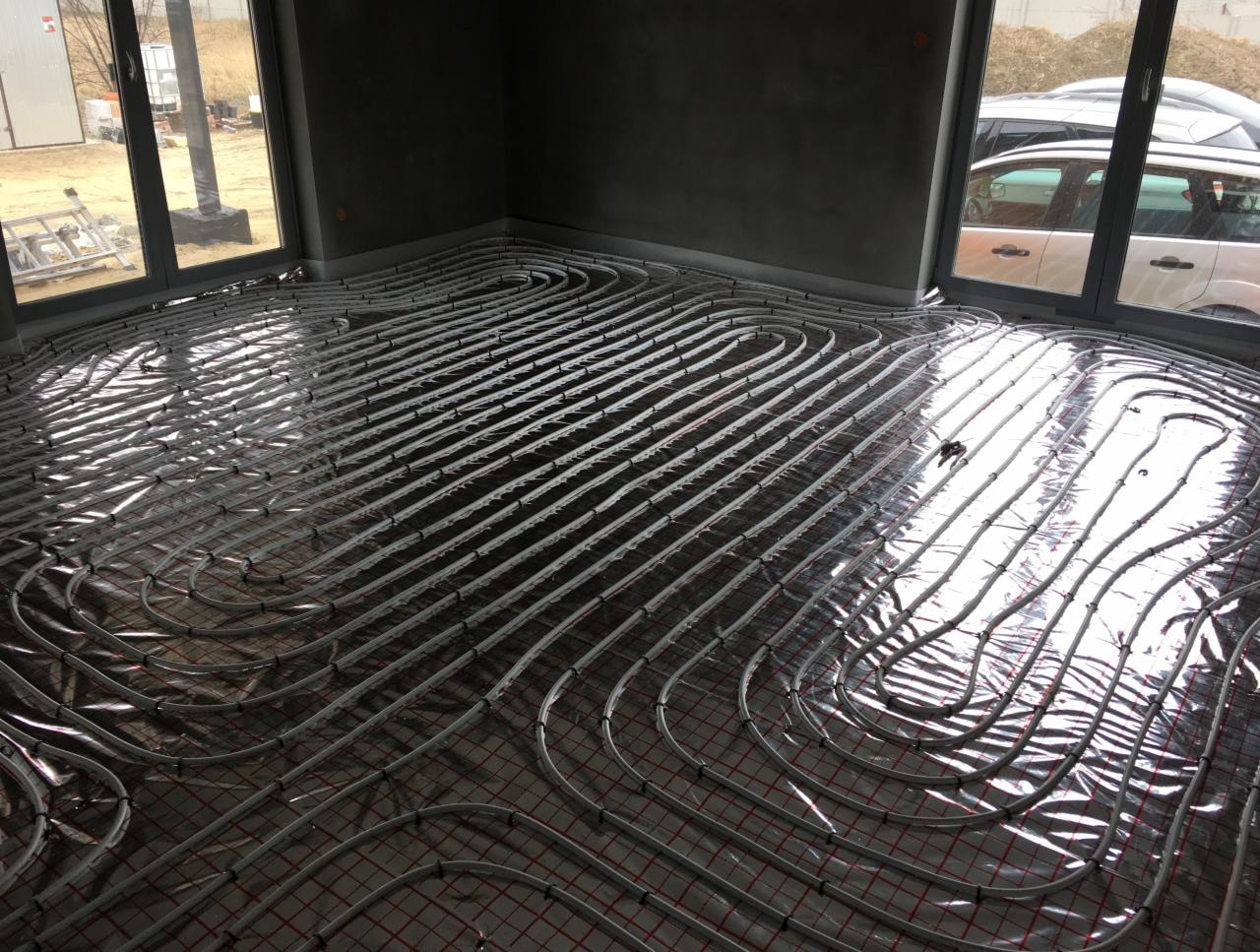 Instalacja ogrzewania podłogowego w surowym pomieszczeniu, widoczne rury grzewcze ułożone na folii i siatce, duże okna z widokiem na zewnątrz.