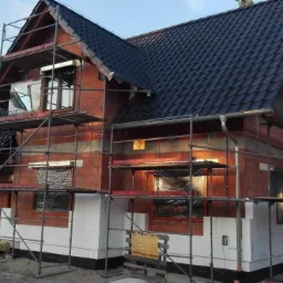 Budowa domu jednorodzinnego w trakcie realizacji: widoczne rusztowania, murowana ściana z czerwonej cegły, ocieplenie styropianem, czarna dachówka i tablica informacyjna budowy.