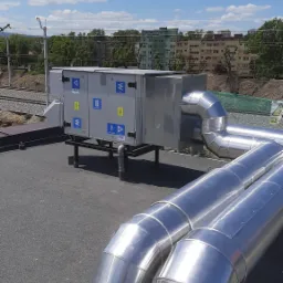 Centrala wentylacyjna z przewodami na dachu budynku, widok na linię kolejową i budynki mieszkalne w tle.