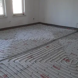 Białe rury grzewcze ułożone spiralnie na szarej macie izolacyjnej z czerwonymi oznaczeniami w surowym pomieszczeniu z dwoma oknami.