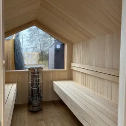 Jasne wnętrze sauny z drewnianymi ławkami, piecem i oknem z widokiem na zewnątrz. Ściany i sufit wyłożone boazerią. Minimalistyczny design, surowy klimat.