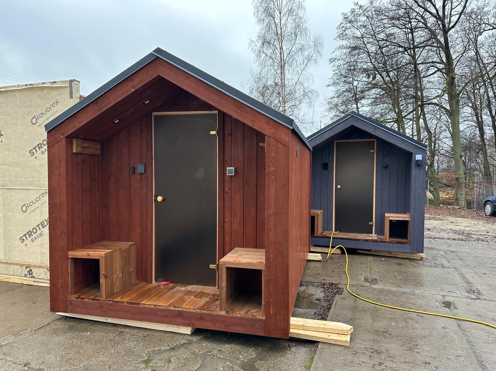 Dwie małe, drewniane sauny ogrodowe z czarnymi drzwiami, ustawione na zewnątrz. Pierwsza w kolorze brązowym, druga szara. Widoczne elementy konstrukcyjne i otoczenie.