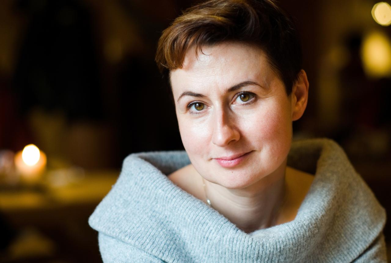 Portret kobiety z krótkimi brązowymi włosami, ubrana w szeroki szary sweter, z delikatnym uśmiechem i ciepłym oświetleniem w tle.