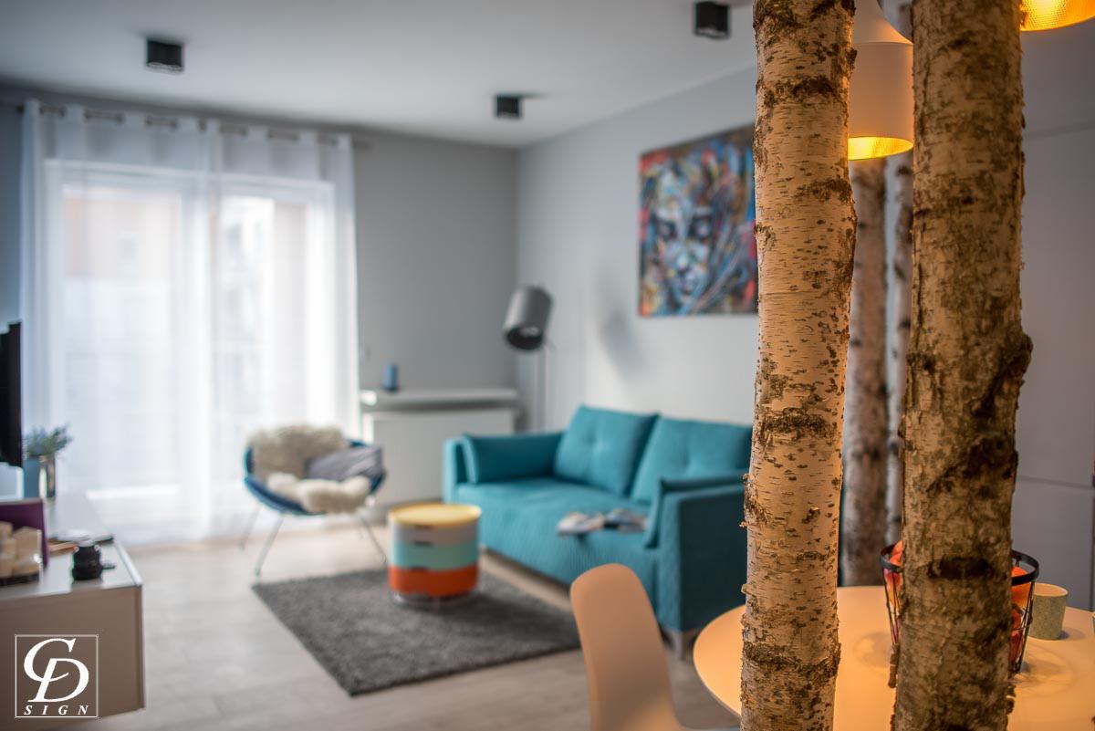 Nowoczesny salon z turkusową sofą, abstrakcyjnym obrazem i dekoracyjnymi pniakami brzozy, oświetlonymi od wewnątrz, tworzącymi element oddzielający przestrzeń.