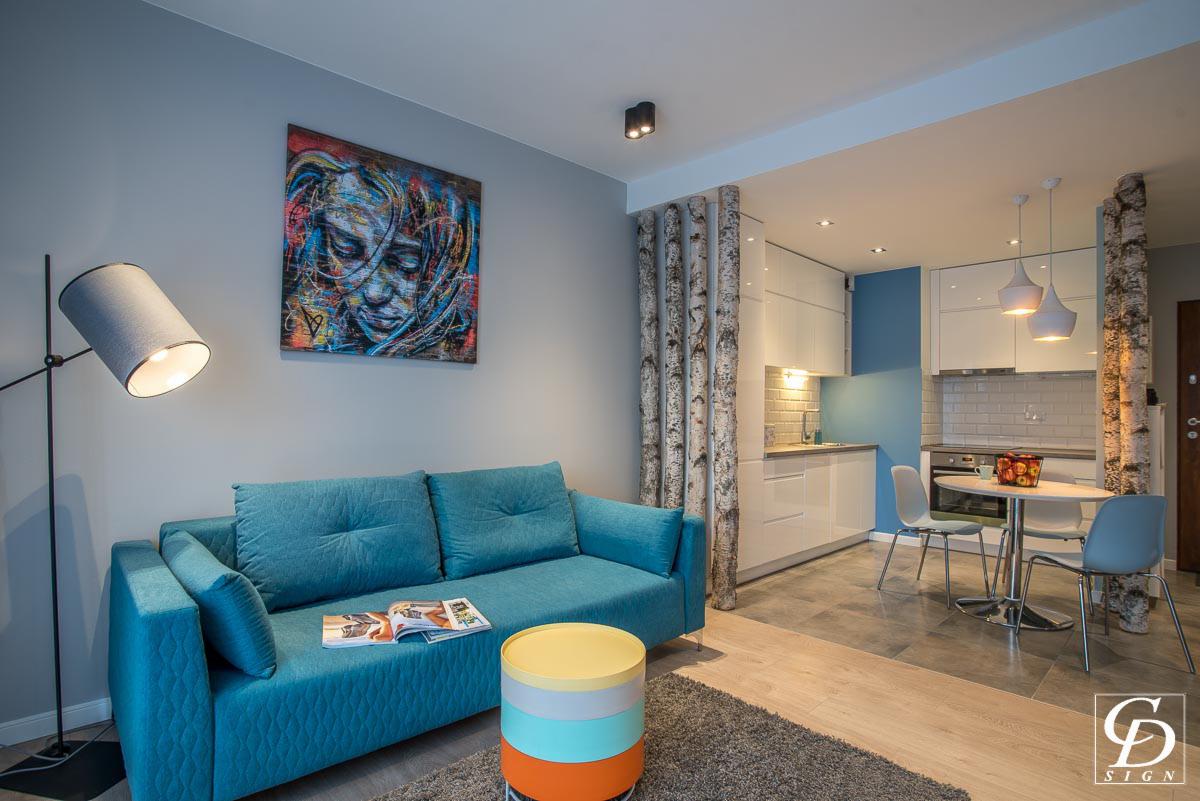 Turkusowa sofa w salonie z obrazem na ścianie i dekoracyjnymi brzozowymi pniami oddzielającymi kuchnię, na stoliku leży otwarta gazeta.