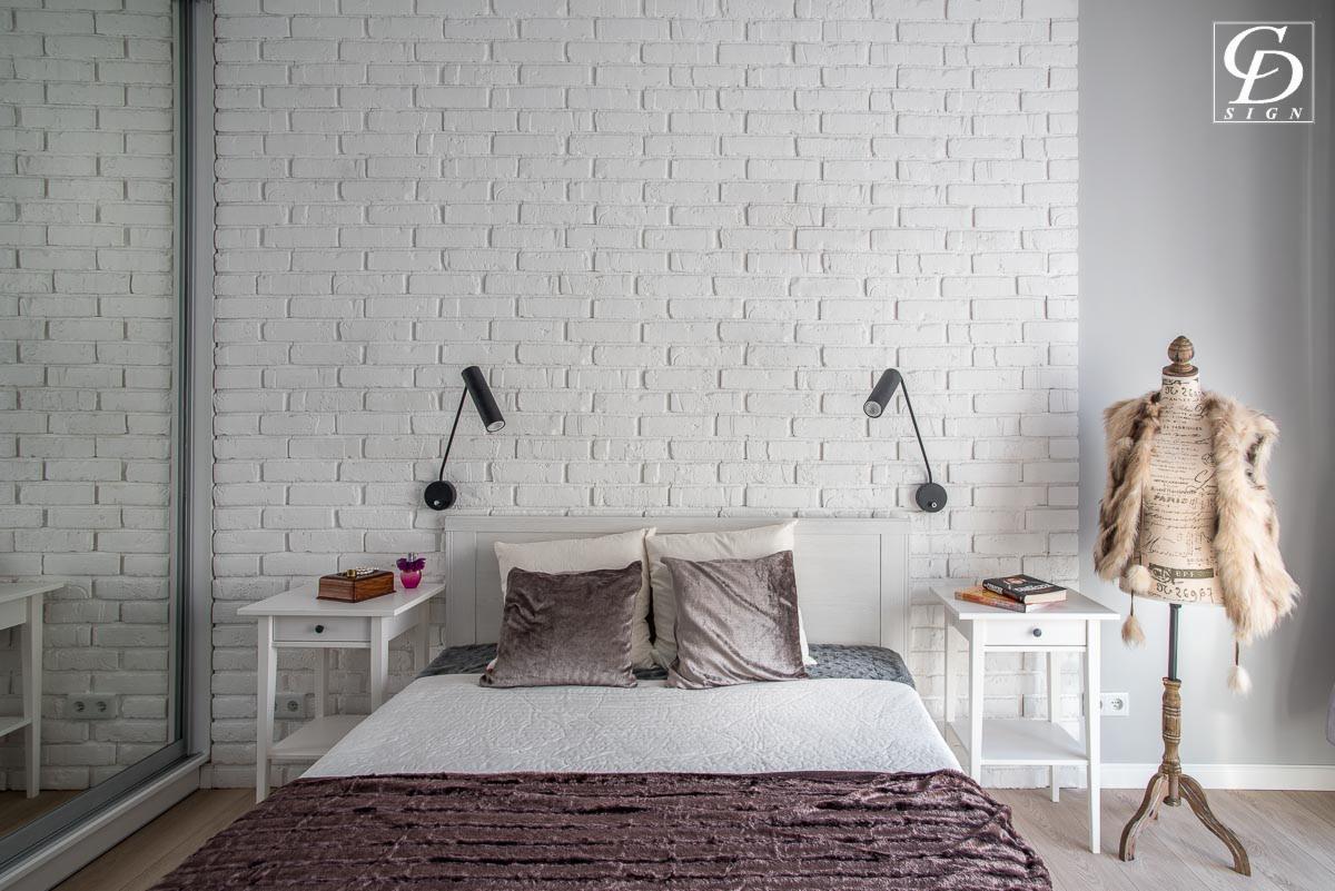 Nowoczesna sypialnia z białą ceglaną ścianą, lampkami nocnymi nad białym łóżkiem z aksamitnymi poduszkami i narzutą w odcieniach fioletu, manekin z futrzaną kamizelką w rogu.