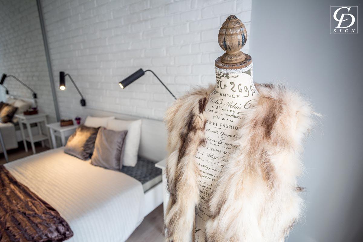 Jasna sypialnia z białymi cegłami na ścianie, łóżkiem z szarymi poduszkami i lampami ściennymi, na pierwszym planie manekin krawiecki z futrzanym szalem.