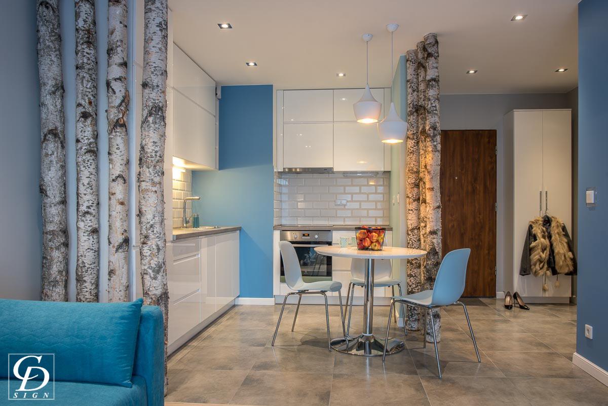 Jasna kuchnia z białymi szafkami, niebieską ścianą i elementami dekoracyjnymi z kory brzozowej, okrągły stół z owocami, krzesła w pastelowych kolorach, widok na przedpokój z szafą i kurtką.