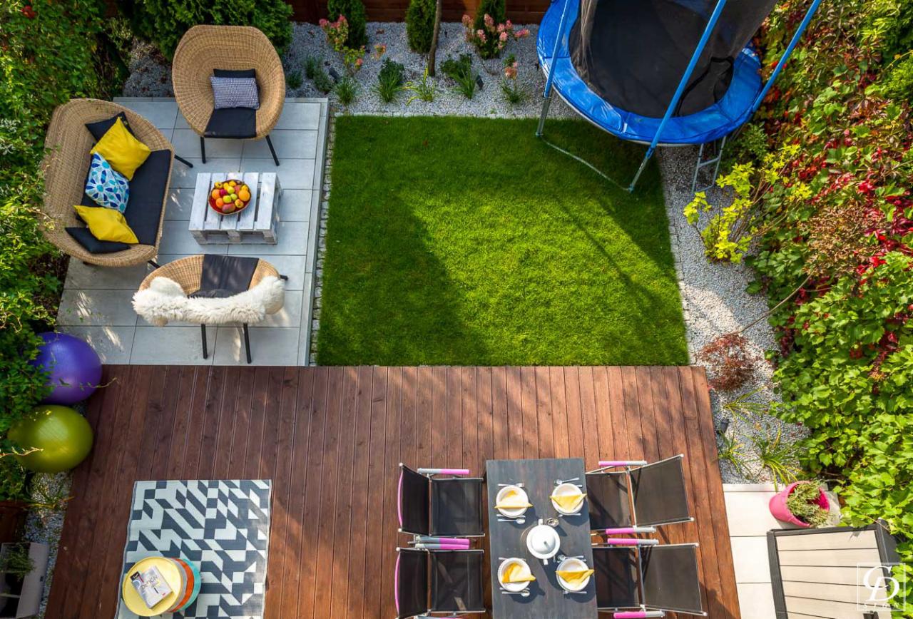Widok z góry na starannie urządzony ogród z drewnianym tarasem, trawnikiem, trampoliną, meblami ogrodowymi z wikliny, stołem z zastawą i dekoracyjnymi roślinami.