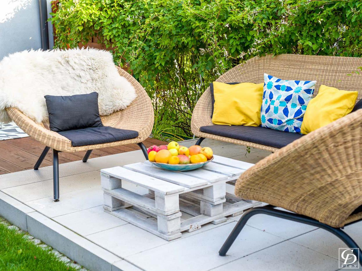 Zewnętrzny kącik wypoczynkowy z wiklinowymi meblami: fotel z owczą skórą, sofa z kolorowymi poduszkami i stolik z palet z misą owoców, na tle zieleni.