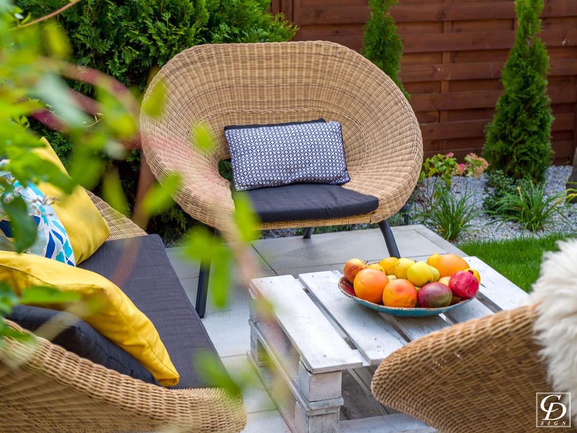 Wygodny rattanowy fotel z poduszką w geometryczny wzór na tarasie, obok stolik z palet z owocami w misie, w tle zadbany ogród z tujami.