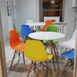 Pok&oacute;j terapii grupowej z okrągłymi białymi stolikami i kolorowymi krzesłami (ż&oacute;łty, pomarańczowy, niebieski, zielony i biały) widziany zza otwartych drzwi.