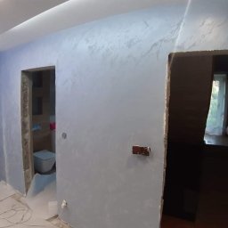 Dariusz - Ściana w odcieniach szarości z efektem dekoracyjnym, widoczne przygotowane otwory drzwiowe i fragment toalety w tle, podłoga zabezpieczona folią malarską.