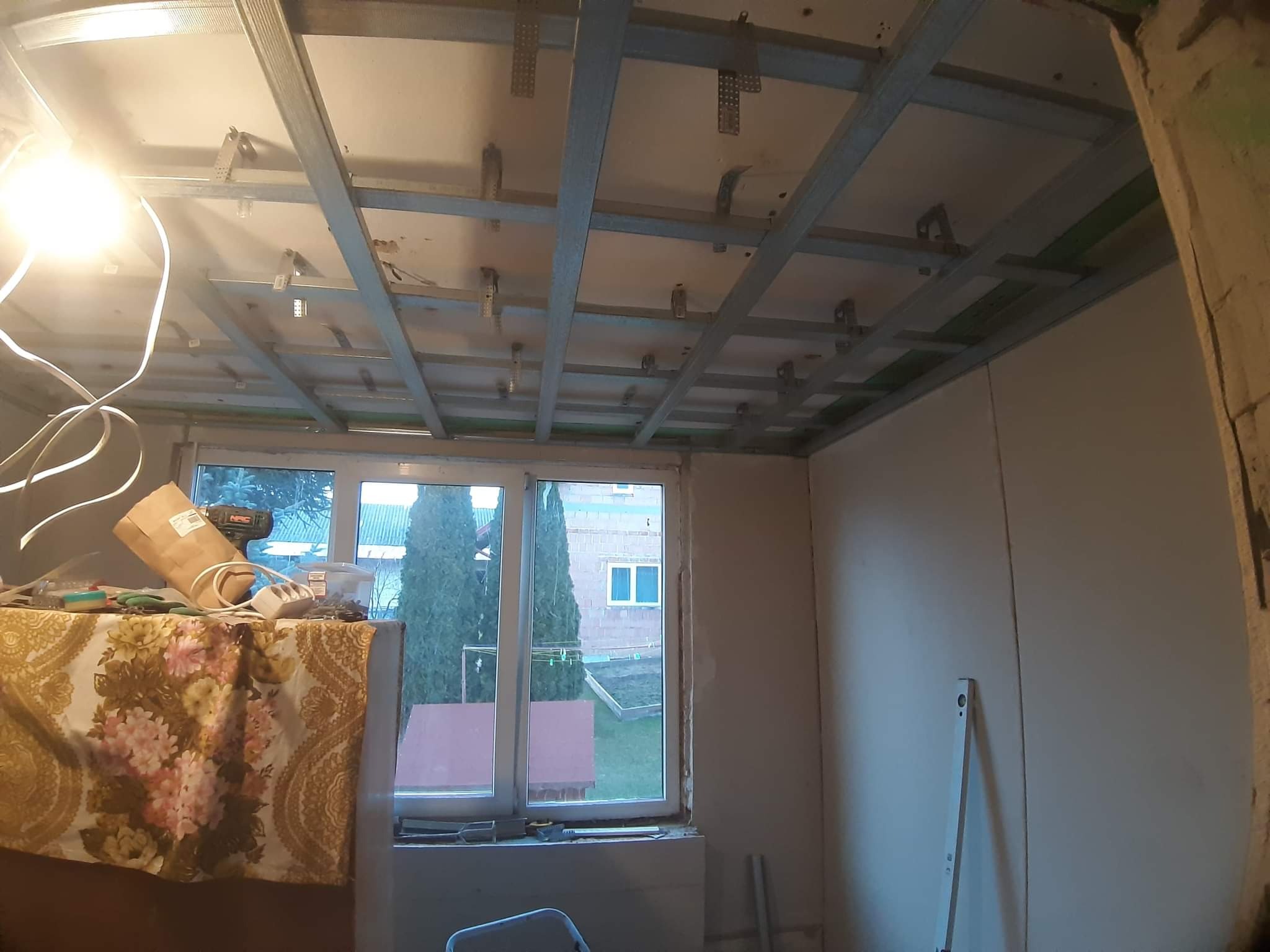 Wnętrze w trakcie montażu sufitu podwieszanego z płyt karton gips, widoczna metalowa konstrukcja rusztu, częściowo zamontowane płyty oraz narzędzia na stole przy oknie.
