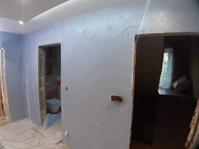 Ściana w odcieniach szarości z efektem dekoracyjnym, widoczne przygotowane otwory drzwiowe i fragment toalety w tle, podłoga zabezpieczona folią malarską.