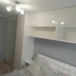 Biała szafa wnękowa z lustrzanymi drzwiami i półkami nad łóżkiem w sypialni, oświetlona trzypunktową lampą sufitową.