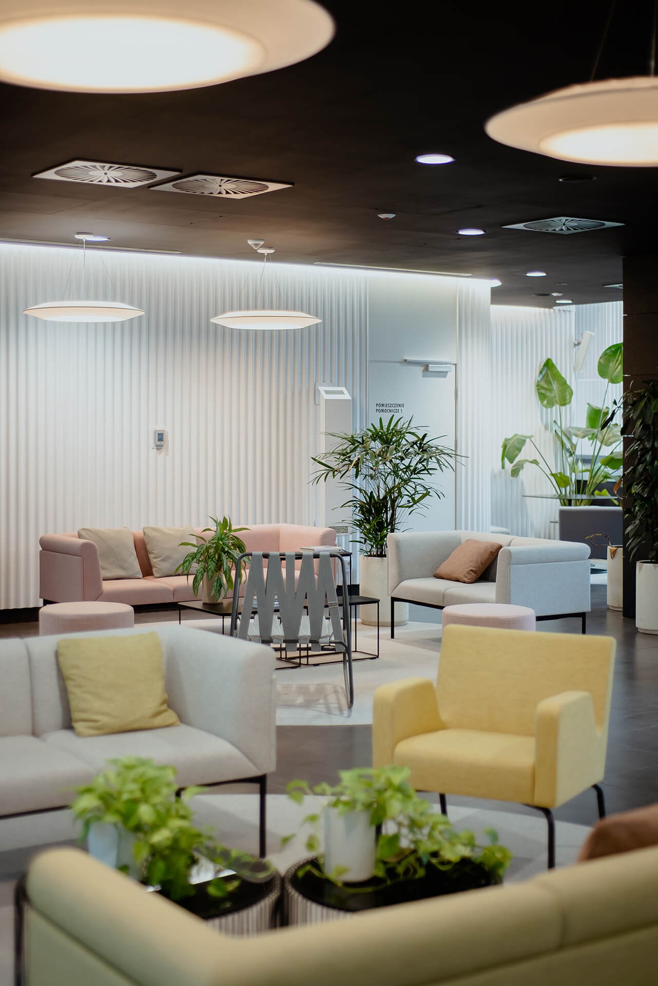 Hol wejściowy biura w stylu retro: pastelowe sofy, geometryczny stolik kawowy, rośliny doniczkowe i oświetlenie dyskowe.