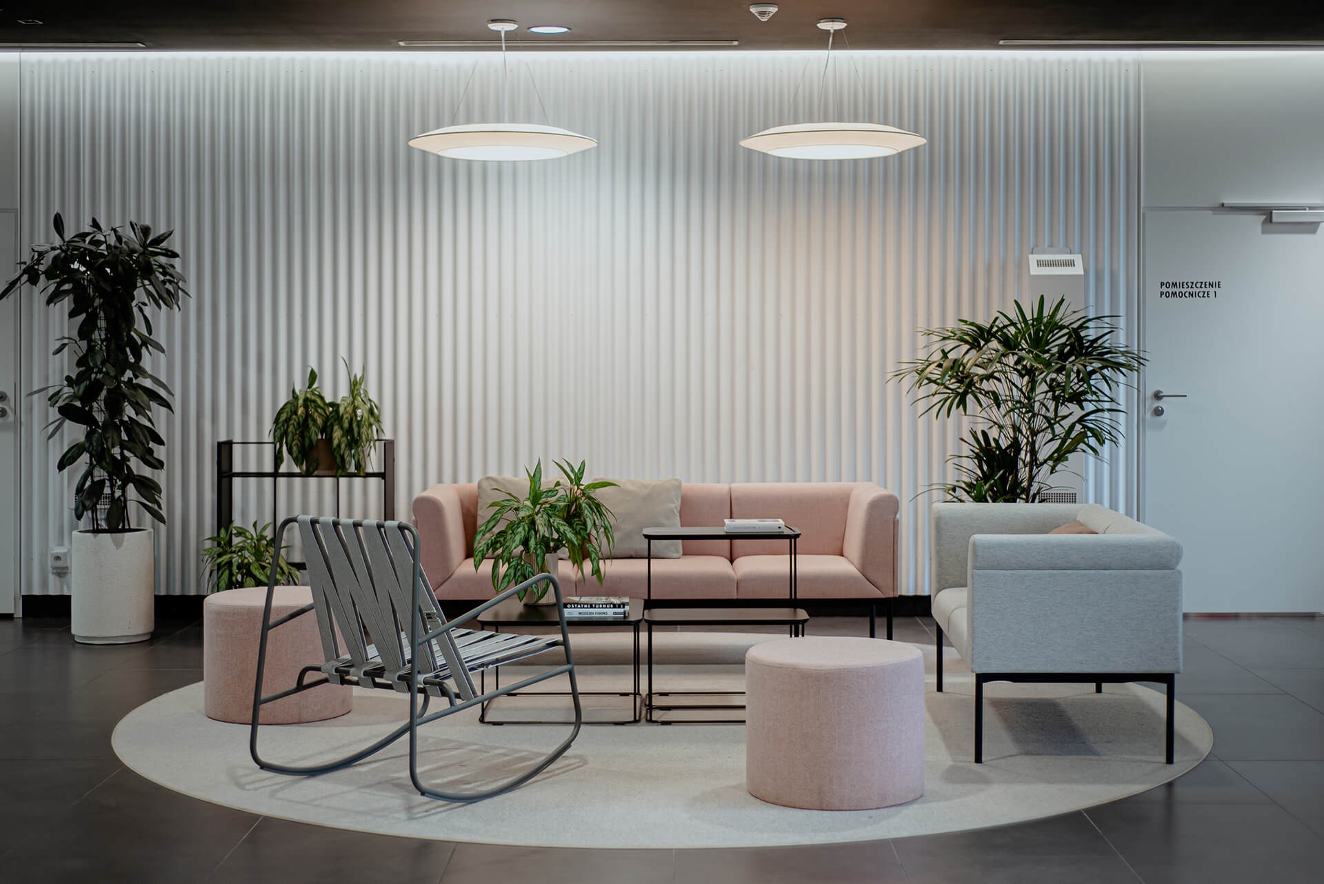 Hol wejściowy w stylu retro: różowa sofa i pufy, szary fotel bujany, nowoczesne lampy wiszące, rośliny doniczkowe na tle białej, żłobkowanej ściany.