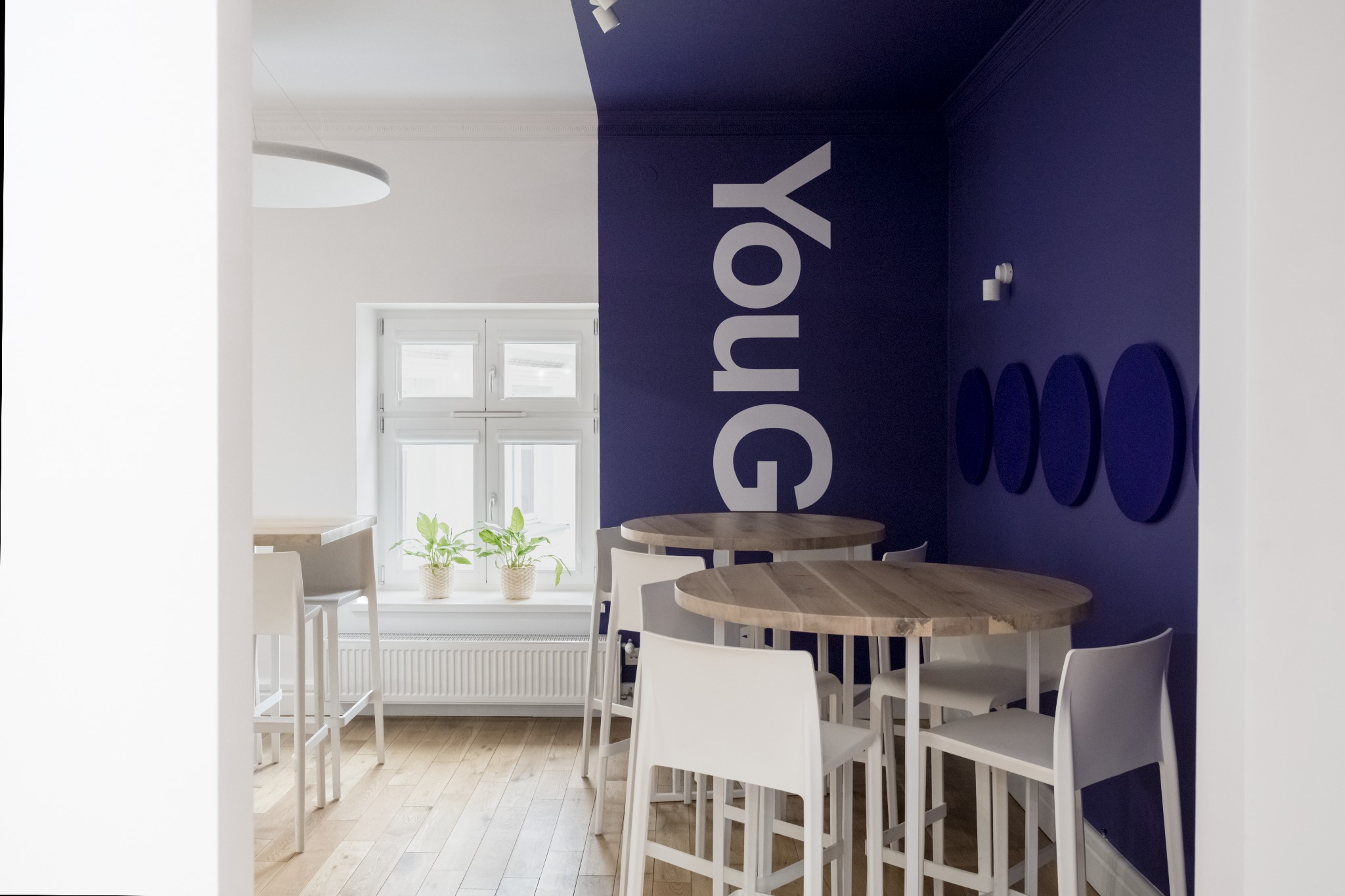 Jasne biuro z drewnianymi stolikami barowymi i białymi krzesłami hokerami, na ścianie logo YouGov i dekoracyjne niebieskie panele akustyczne.