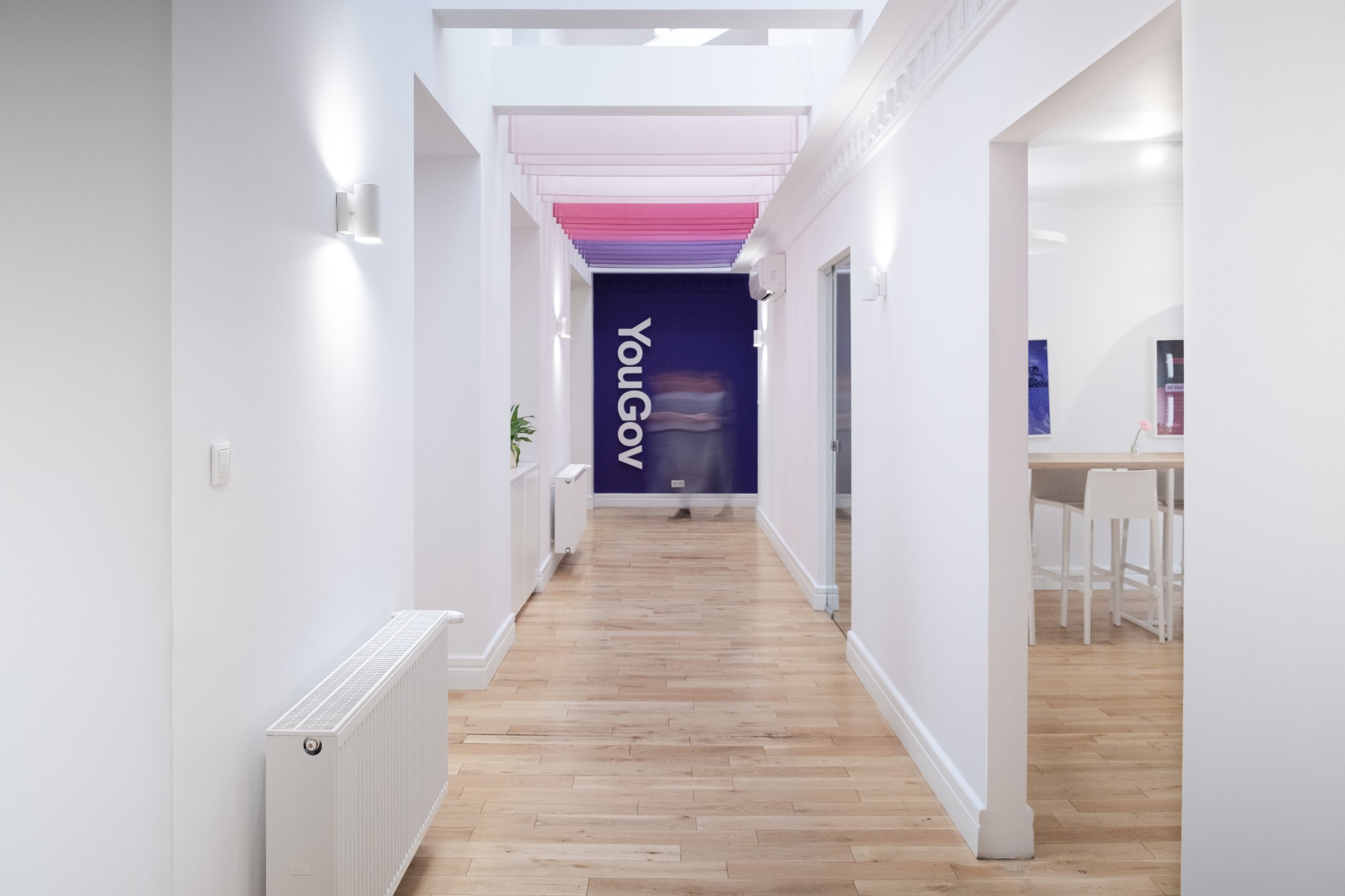 Jasny korytarz biurowy z drewnianą podłogą, białymi ścianami, grzejnikami i logo YouGov na ścianie. W tle widoczne biurko i krzesła.