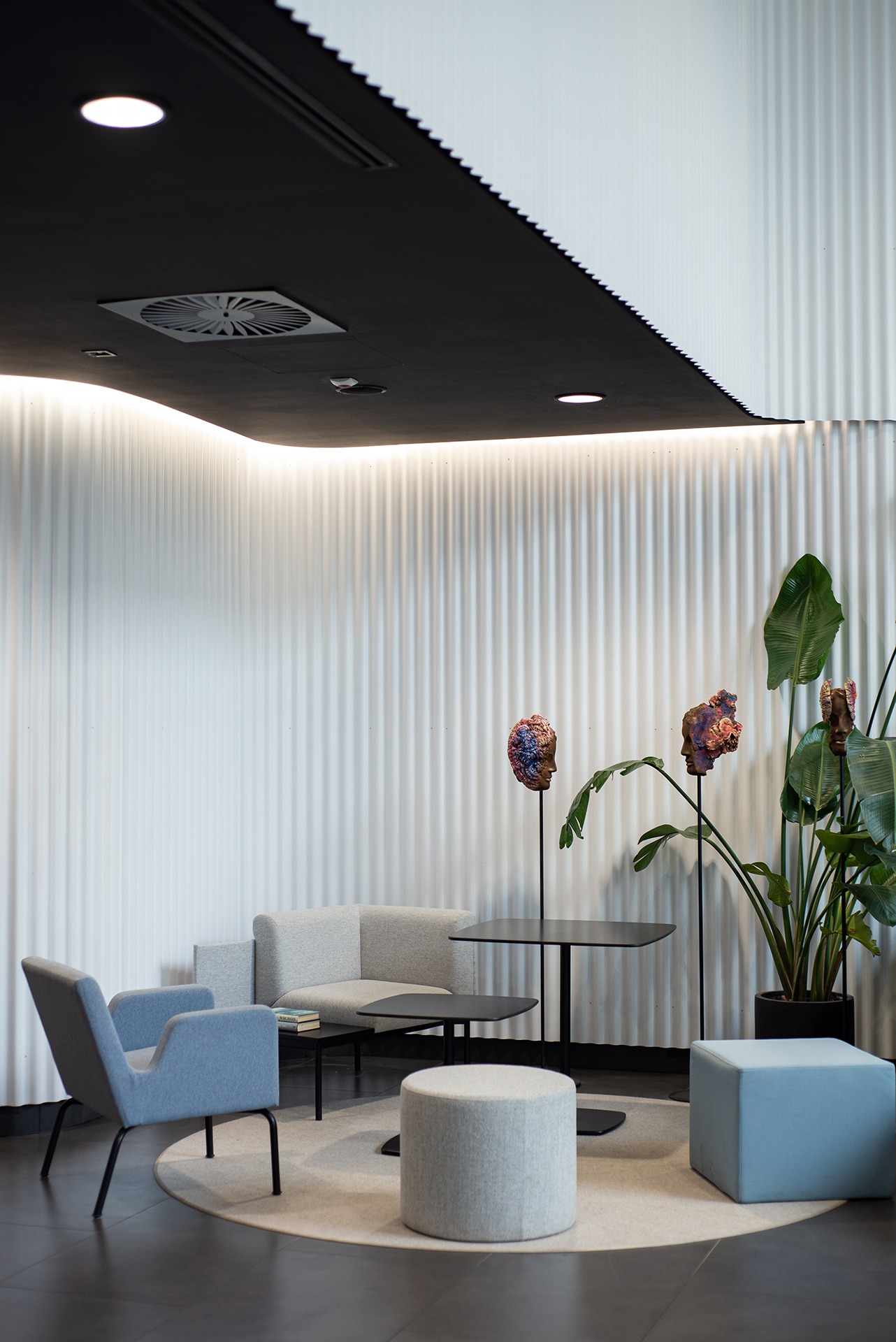 Nowoczesna przestrzeń biurowa z szarymi meblami wypoczynkowymi, czarnymi stolikami, rzeźbami głów na stojakach i rośliną doniczkową na tle białej, prążkowanej ściany i czarnego sufitu...