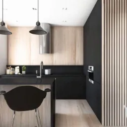 Nowoczesna kuchnia z czarnymi szafkami, drewnianymi panelami ściennymi i wyspą z czarnym blatem oraz designerskimi krzesłami barowymi, oświetlona trzema wiszącymi lampami w stylu industrialnym.