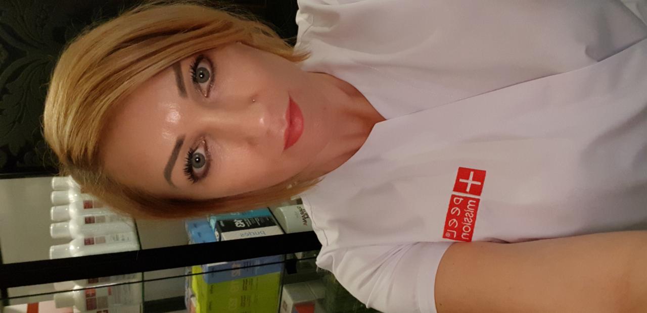 Portret kobiety w białym uniformie z logo firmy, z delikatnym makijażem, w tle półki z kosmetykami.