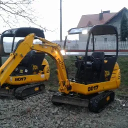 Dwa żółte mini-koparki JCB 8017 na żwirowym podłożu, widoczne oświetlenie robocze, w tle budynek mieszkalny i drzewa.