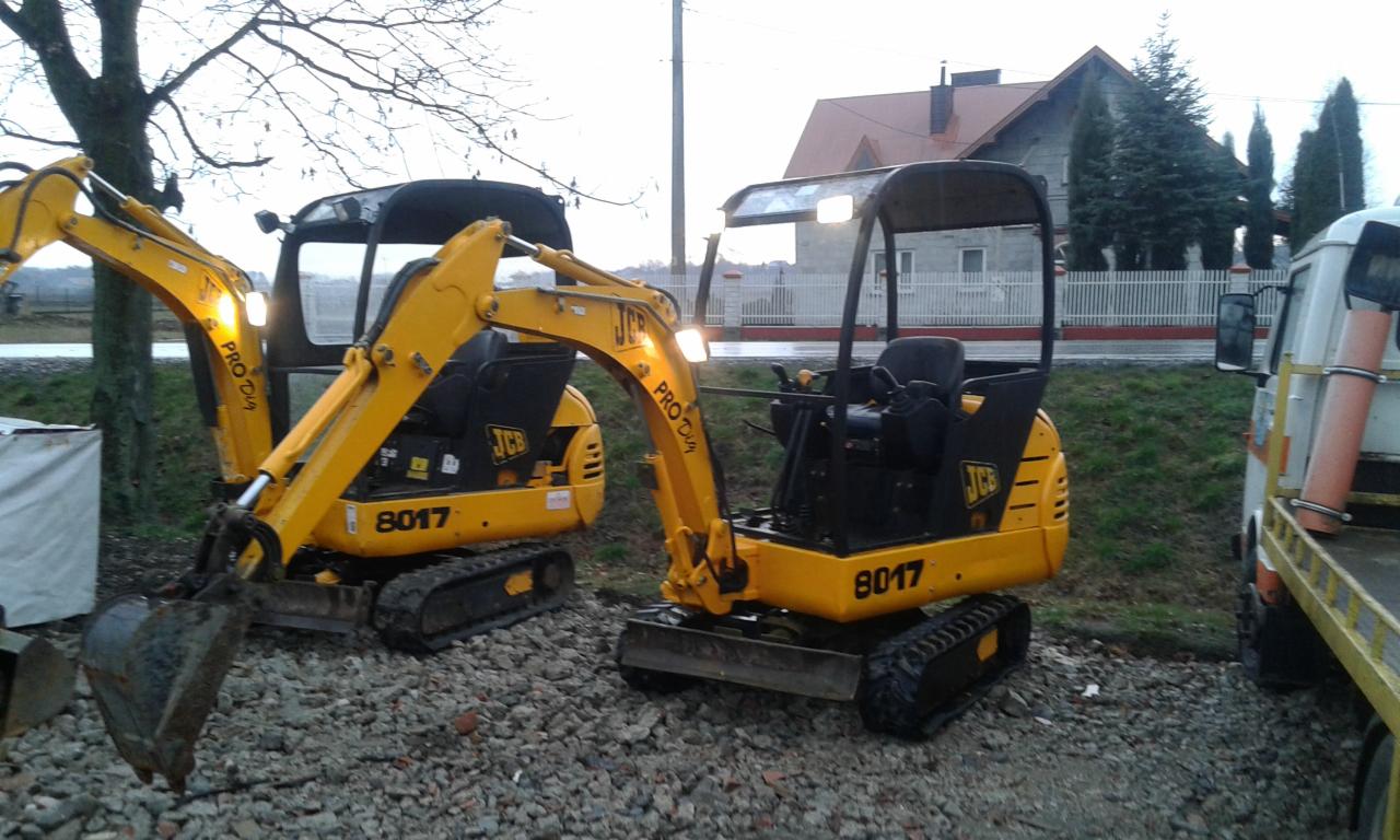 Dwa żółte mini-koparki JCB 8017 na żwirowym podłożu, widoczne oświetlenie robocze, w tle budynek mieszkalny i drzewa.