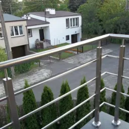 Błyszcząca, metalowa balustrada balkonowa z widokiem na posesję z dwoma domami, zielonymi tujami i zaparkowanym samochodem.