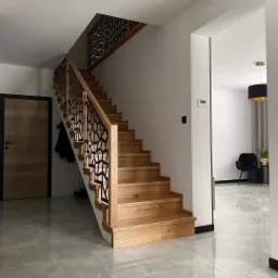 Nowoczesne schody drewniane z ażurową balustradą metalową w jasnym wnętrzu domu z szarymi płytkami podłogowymi.