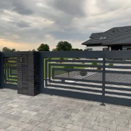 Nowoczesna brama wjazdowa z elementami dekoracyjnymi w kształcie kwadratów i perforowaną siatką, osadzona na słupkach z ciemnej cegły klinkierowej, na tle domu z szarą dachówką i pochmurnego nieba.