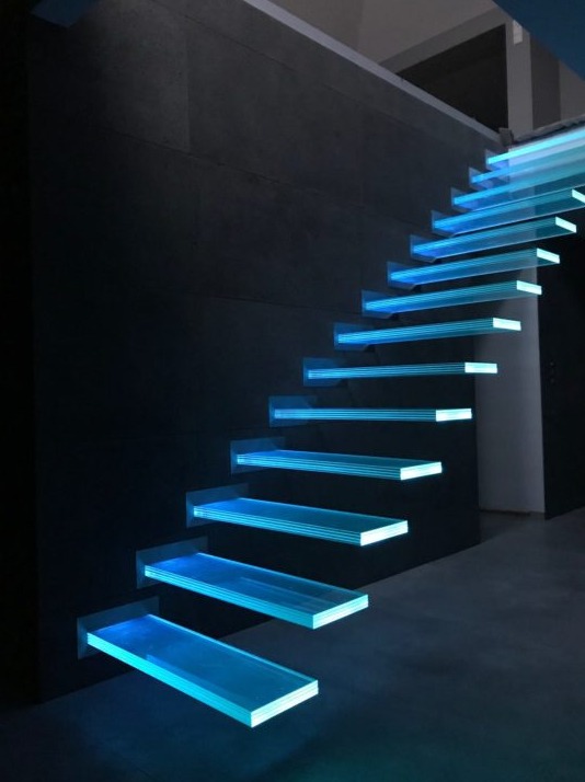 Nowoczesne schody z podświetlanymi na niebiesko, szklanymi stopniami, zamontowane przy ciemnej ścianie w Radomiu.