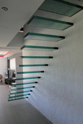 Szklane schody wspornikowe z czarnymi mocowaniami na tle białej ściany z delikatnym wzorem, widoczne z boku, w nowoczesnym wnętrzu.