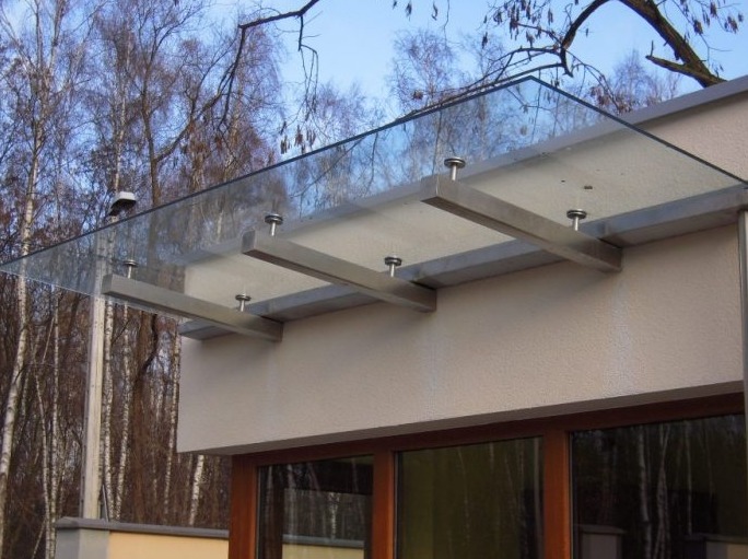 Szklany daszek nad wejściem, wsparty na metalowych wspornikach, widok z dołu na tle drzew bez liści.